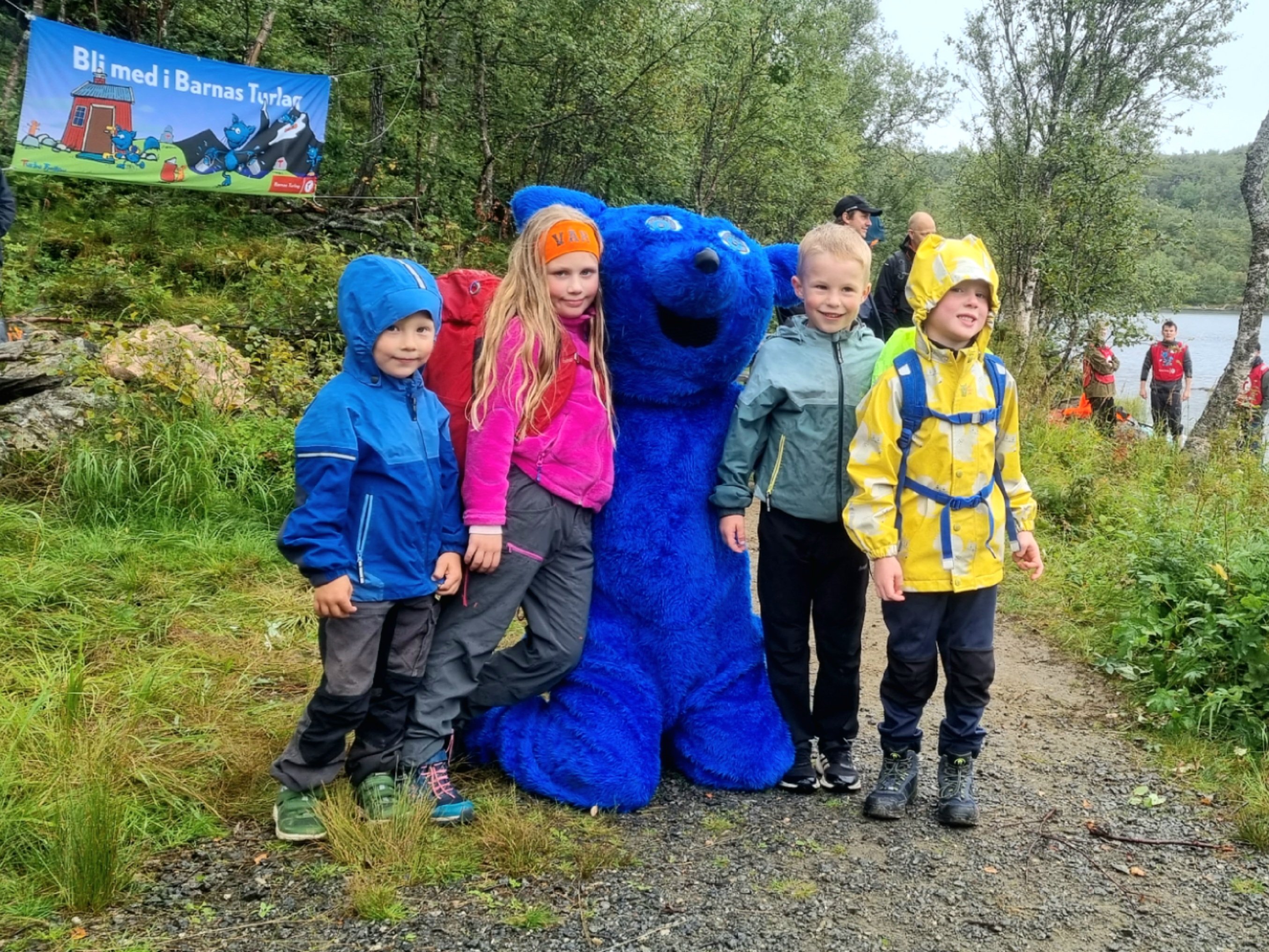 Fire barn står ved siden av en stor, blå rev som sitter på kne. De er ute i skogen. Barna har regntøy i ulike farger.
